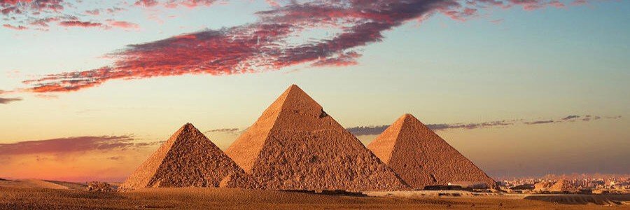 Panoramic view of the three main stone pyramids of Giza standing tall in the Egyptian desert sand under a vibrant twilight sky filled with long streaking clouds illuminated in bright shades of pink, orange, and deep purple during sunset.