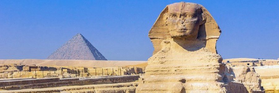 A clear, sunny view of the Great Sphinx of Giza with the peak of a pyramid standing in the background against a bright blue sky.