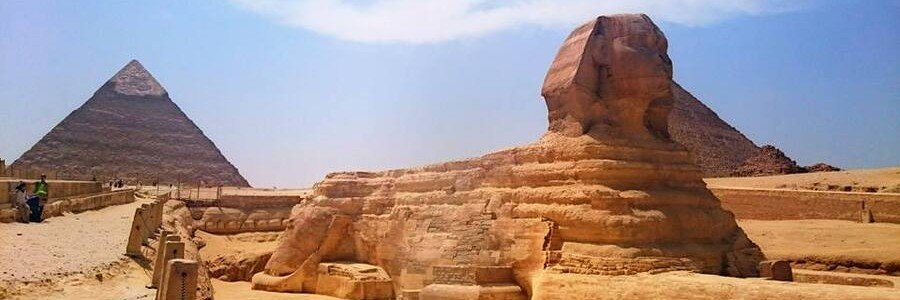 The Great Sphinx of Giza shown in profile view with the ancient Pyramid of Khafre rising in the background under a bright, hazy sky.