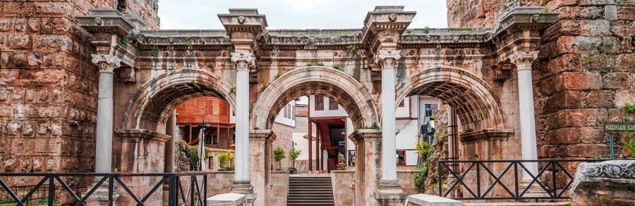 hadrians-gate-antalya-old-town-entrance | Beach Travel Destinations A panoramic front view of the white marble Hadrian's Gate in Antalya, showcasing three symmetrical Roman arches with ornate Corinthian columns, set against a backdrop of historic red-brick towers and lush green hills.