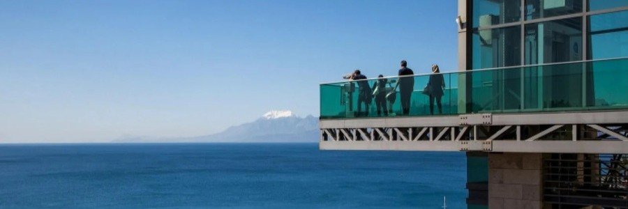 kaleici-panoramic-elevator-antalya-view | Beach Travel Destinations A wide-angle shot of tourists standing on the glass-railed observation deck of the Kaleiçi Panoramic Elevator in Antalya, overlooking the deep blue Mediterranean Sea and the snow-capped Taurus Mountains in the far distance.