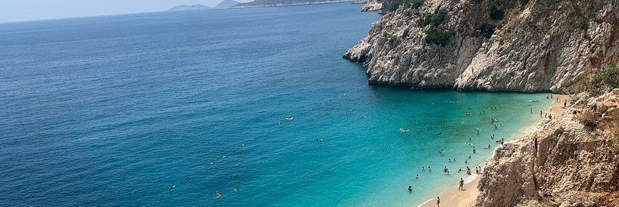 kaputas-beach-antalya-turquoise-coast | Beach Travel Destinations A high-angle panoramic view of Kaputaş Beach, featuring brilliant turquoise Mediterranean water meeting a golden sandy cove, bordered by massive limestone cliffs with people swimming in the clear bay.