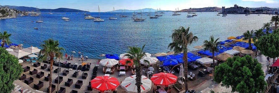 Panoramic view of Kumbahçe Beach in Bodrum featuring colorful beach umbrellas, outdoor restaurant seating, several boats anchored in the blue bay, and Bodrum Castle on the horizon.
