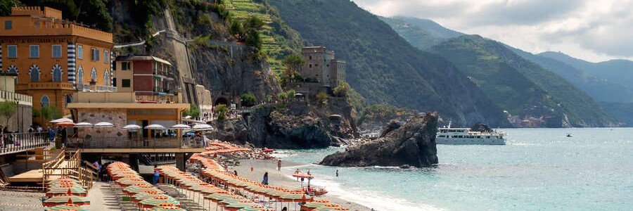 Monterosso al Mare beach in Cinque Terre featuring orange umbrellas, coastal buildings, and a ferry on the turquoise water near La Spezia Port.