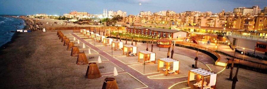 High-angle twilight view of the Marom Port Said Resort beach showing illuminated luxury cabanas, a sandy shoreline bordering the Mediterranean Sea, and the resort property glowing with warm lights.