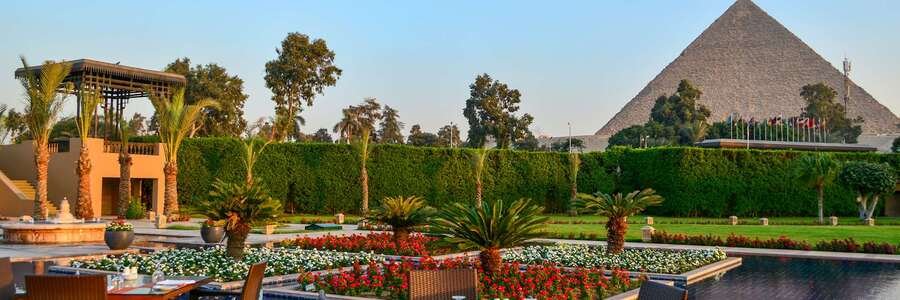 A view from the lush garden terrace of the Marriott Mena House, showing a large pool, flower beds, and the Great Pyramid of Giza in the background.