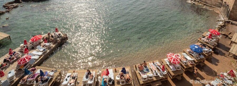 mermerli-beach-antalya-old-town-summer | Beach Travel Destinations A high-angle view of Mermerli Beach in Antalya's Old Town, featuring several wooden sunbathing platforms over the water and a narrow rocky shore lined with dozens of sun loungers under vibrant red and white umbrellas, with shimmering turquoise Mediterranean water in the foreground.