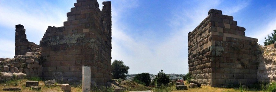 Two large, ancient stone towers of the Myndos Gate in Bodrum, Turkey, with a white information plaque in the foreground and a clear blue sky above.
