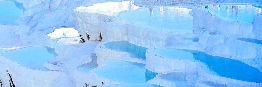 pamukkale-travertine-thermal-pools-turkey | Beach Travel Destinations A wide panoramic view of the brilliant white travertine terraces at Pamukkale, Turkey, featuring numerous stepped, shallow pools filled with clear turquoise thermal water under a bright sky.