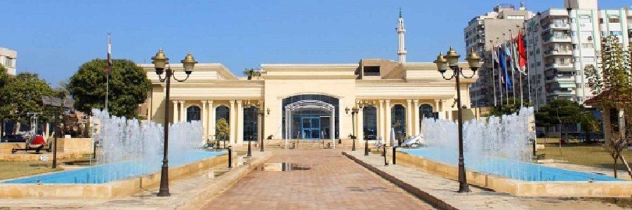 The exterior entrance of the Port Said Military Museum featuring long blue fountain pools spraying water in front of a symmetrical beige building under a clear blue sky.