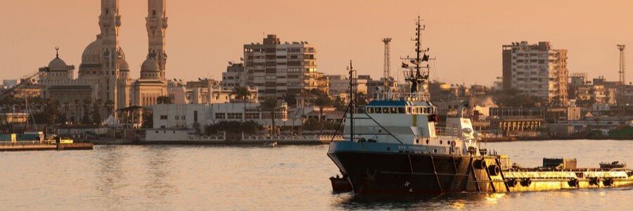 A view across the Suez Canal at sunset showing a large utility vessel in the foreground water and the twin minarets of the Grand Mosque rising above the Port Said city skyline in Egypt.