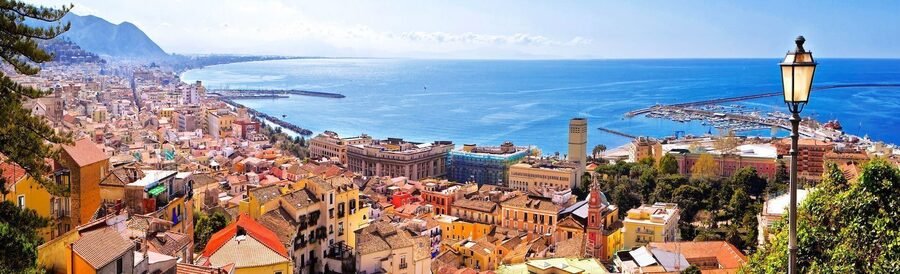 High-angle view of colorful pastel buildings, terracotta roofs, and the curved coastline of Salerno meeting the Tyrrhenian Sea.