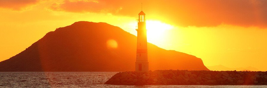  Silhouette of the Turgutreis lighthouse on a rocky coastline at sunset with a large island and golden orange sky in the background.