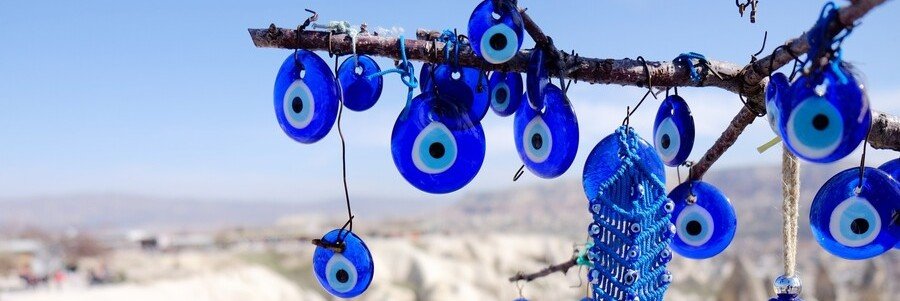 turkish-evil-eye-amulets-cappadocia | Beach Travel Destinations A close-up shot of numerous bright blue glass Nazar amulets (evil eye charms) of various sizes hanging from a weathered tree branch, with the blurred, beige-colored rocky landscape of Cappadocia under a clear blue sky in the background.