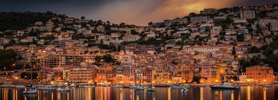 Villefranche-sur-Mer harbor at dusk with glowing lights reflecting on calm water and hillside homes on the French Riviera
