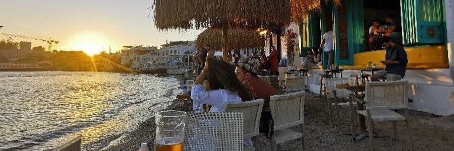  Patrons relaxing under thatched umbrellas at Yula Bodrum beach bar during a golden sunset overlooking the Aegean Sea.