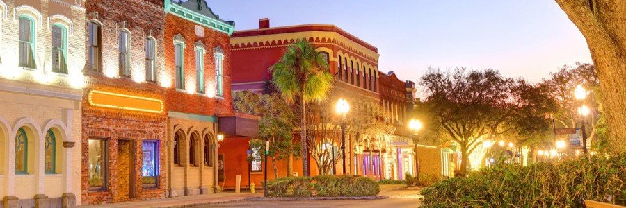 Historic downtown street at dusk with brick storefronts, a palm tree, and warm street lamps glowing along the sidewalk.