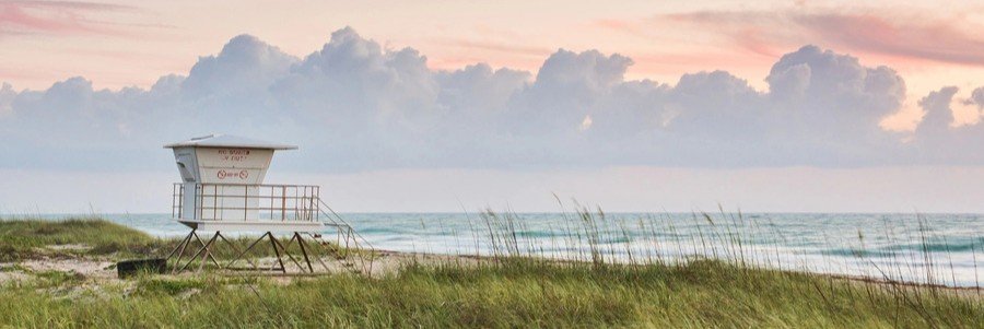 A white beach lifeguard tower stands behind tall grass sea oat dunes facing a calm ocean under a pink tinted sunset sky.