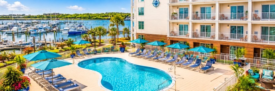 Sparkling turquoise swimming pool surrounded by blue lounge chairs and umbrellas, with a marina full of boats in the background.