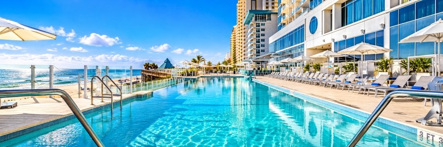 Sparkling oceanfront swimming pool with lounge chairs, white umbrellas, and Atlantic Ocean views at Daytona Grande Resort in Daytona Beach, Florida.