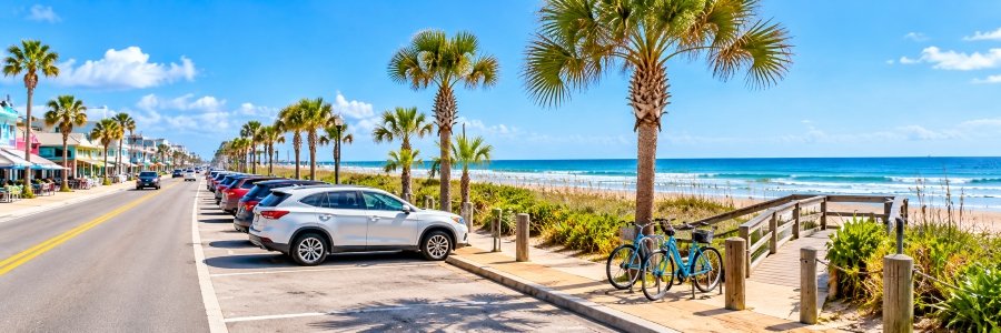 Cars parked along a palm-lined coastal road beside a wooden beach access walkway, with bicycles and ocean views.