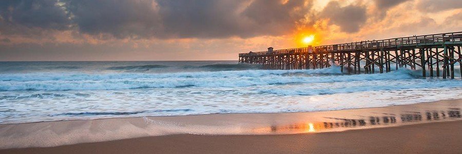 A long wooden fishing pier extends into the Atlantic ocean at sunrise on Flagler Beach, Florida, with waves crashing on the orange coquina sand shoreline.