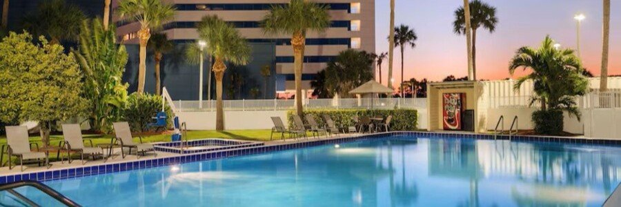 Hotel pool area at dusk with palm trees, lounge chairs, and a vending machine near a modern building.