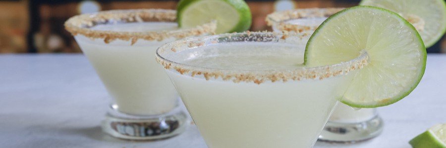 Close-up of three margarita glasses with salted rims and lime wheels as garnishes on a light table.