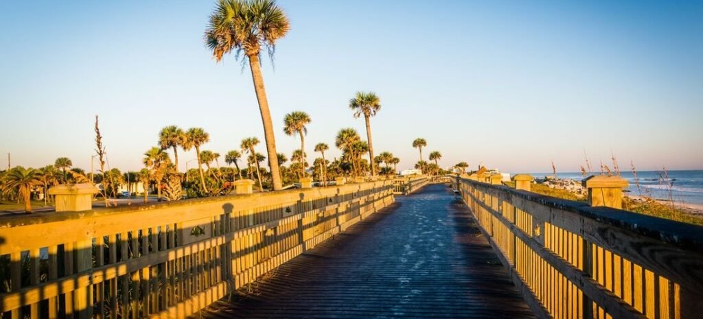 A long wooden oceanfront boardwalk winds past tall palm trees along the scenic coastline of Palm Coast, Florida under a clear blue sky.