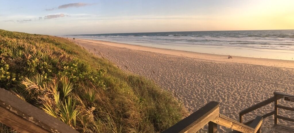 Wooden boardwalk stairs leading down to the wide, untouched sandy shores of Playalinda Beach at sunset.