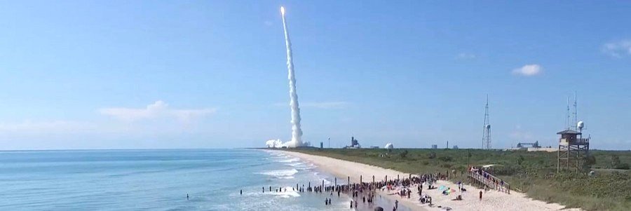 A large crowd gathered on the sandy shore of Playalinda Beach watches an Atlas V rocket launch from Cape Canaveral, leaving a massive white smoke trail in a clear blue sky.