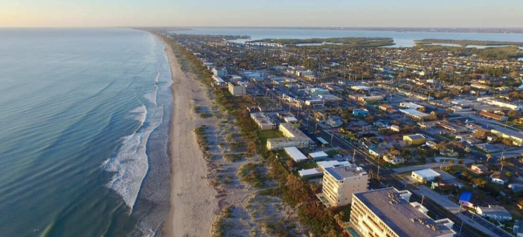 High aerial view looking down the expansive coastline of Satellite Beach, Florida, showing the ocean waves, wide sandy beach, and city buildings.