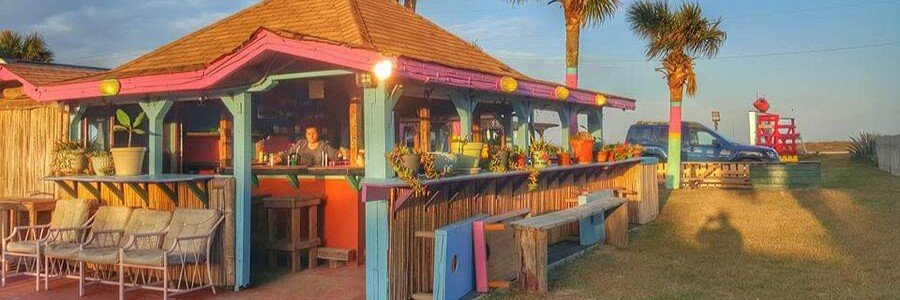 Colorful open-air beach bar with a pink roof, turquoise walls, and palm trees, serving drinks at sunset.