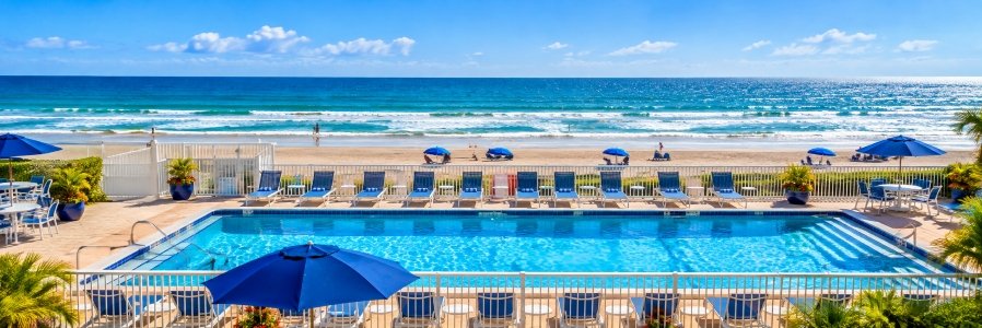 Sparkling beachfront swimming pool with blue lounge chairs, umbrellas, and ocean waves along the sandy shore in Ormond Beach, Florida.