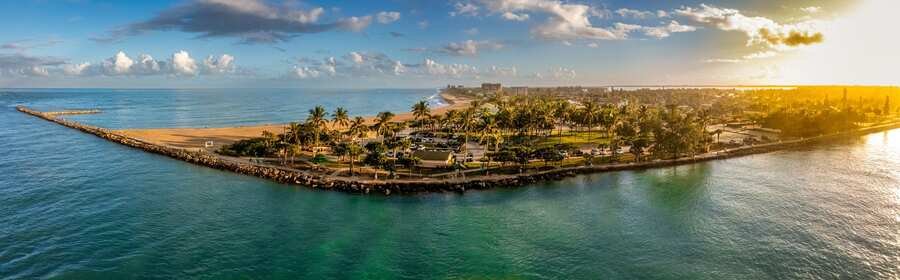 Aerial view of Fort Pierce Inlet with turquoise water, a palm-lined shoreline, sandy beach, and rock jetty