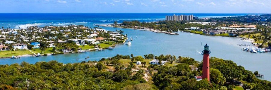 Aerial view of a red lighthouse surrounded by lush green trees next to a bright blue ocean inlet.