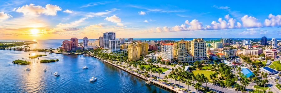 Bright coastal skyline in Boynton Beach, Florida, featuring blue waterfront waters, sailboats, palm-lined streets, and colorful buildings beneath a sunny morning sky.