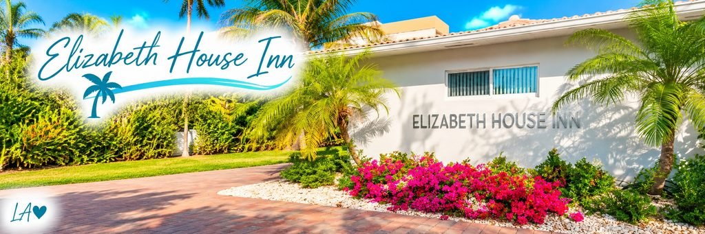 Sunny entrance to Elizabeth House Inn, featuring a white stucco building, tropical palm trees, bright pink flowers, and a brick walkway.