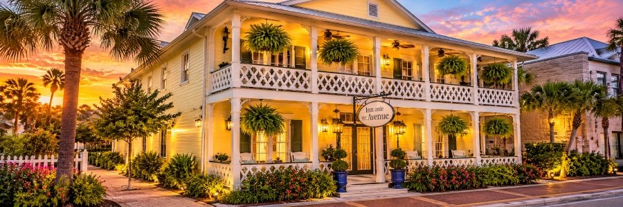 Inn on the Avenue | New Smyrna Beach Charming yellow coastal inn with white verandas, tropical plants, and welcoming porch seating at sunset.