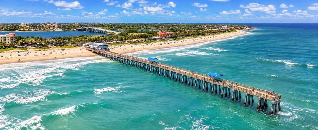 Aerial view of Lake Worth Beach Pier in Florida, stretching into turquoise Atlantic Ocean waters beside sandy beaches and a sunny coastline.