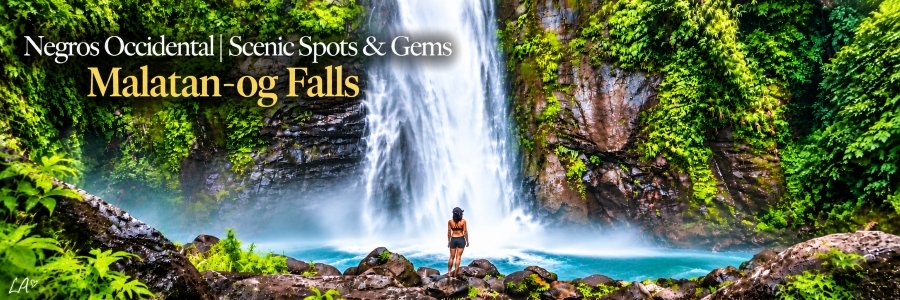 Towering waterfall cascading down a rocky cliff surrounded by lush tropical greenery, with a woman standing on boulders beside a turquoise pool.