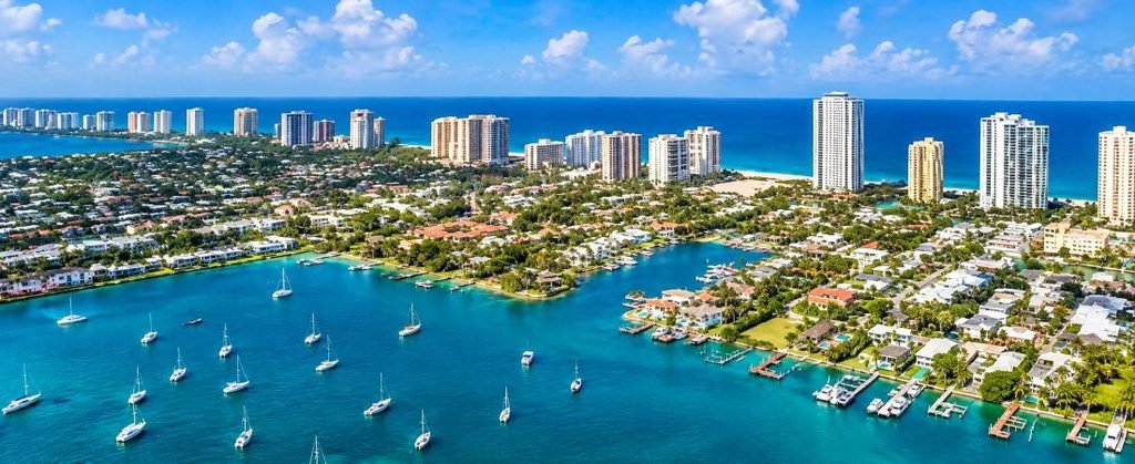 A stunning enhanced aerial view of Palm Beach, Florida, featuring sparkling turquoise waterways, beautiful sailboats, luxury waterfront homes, tropical landscaping, and an impressive Atlantic Ocean skyline.