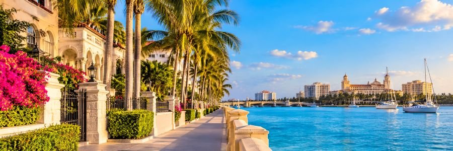 Elegant Mediterranean-style waterfront estates surrounded by tropical palm trees, vibrant pink flowers, and sparkling turquoise water with luxury yachts.