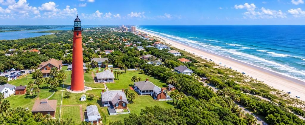 Ponce Inlet | Amazing Beaches & Surfing in Florida Ponce Inlet Florida aerial view featuring the historic red lighthouse, lush coastal greenery, sandy Atlantic beaches, and sparkling ocean waves.