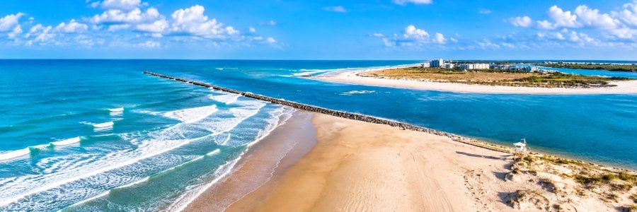 Ponce Inlet Florida aerial view featuring a long rock jetty, golden sandy beach, turquoise Atlantic waves, and scenic coastal waterways.
