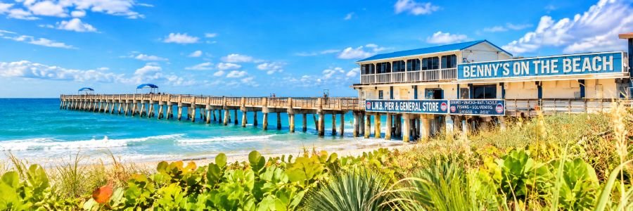 Sunny view of Lake Worth Beach Pier and Benny's on the Beach in Lake Worth Florida, with turquoise ocean water, blue skies, and coastal greenery.