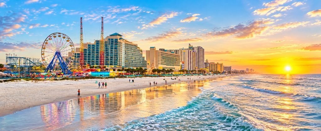 Daytona Beach Shores travel guide hero featuring a golden Atlantic sunrise, beachfront hotels, colorful amusement rides, and sparkling ocean waves.